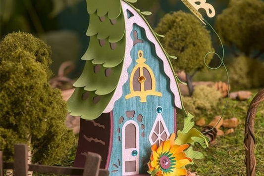 Butterfly Fairy House 3D - craftkindle: A whimsical teal-colored paper craft fairy house with white trim, featuring delicate door details, surrounded by mossy green elements and a bright sunflower accent. The house stands against a natural woodland-inspired background.